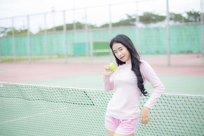 Portrait of young woman holding ball while standing against net on court