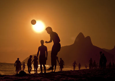 Silhouette friends playing soccer at beach against orange sky during sunset
