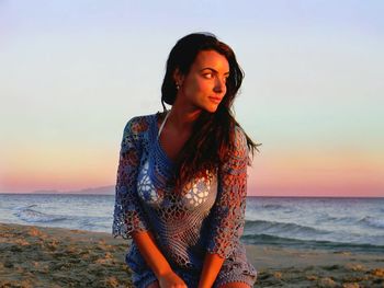 Beautiful young woman looking away while relaxing on shore at beach during sunset