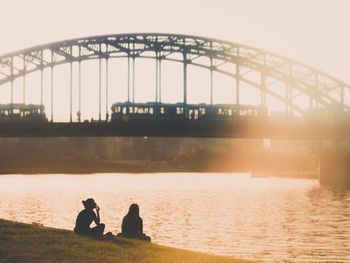 Silhouette people sitting on bridge over river against sky