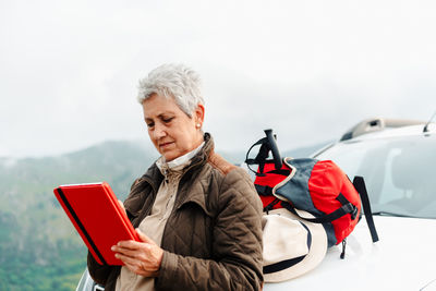 Elderly female traveler with short gray hair leaning on car and browsing tablet during road trip in countryside