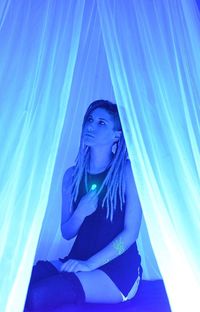 Young woman looking away while sitting against illuminated blue curtain