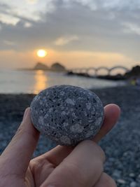 Close-up of hand holding pebble against sea during sunset