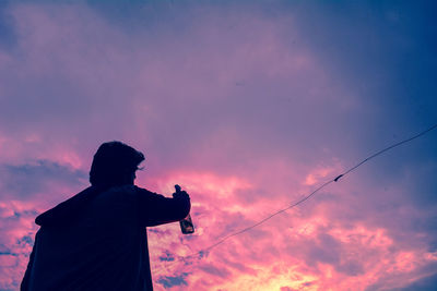 Low angle view of man holding bottle looking at dramatic sky during sunset