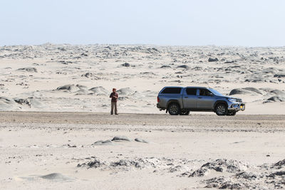 Man standing by car on sand in desert against clear sky