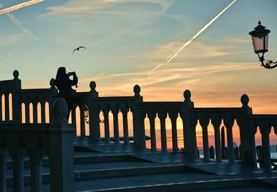 Silhouette woman photographing vapor trail by sea against sky during sunset