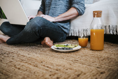 Young man working on laptop while sitting by juice and fruit plate on floor at home