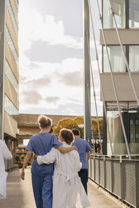 Rear view of healthcare workers with arms around each other walking towards hospital during sunset