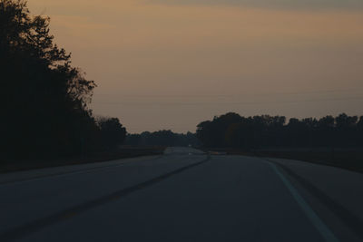Road by silhouette trees against sky at sunset