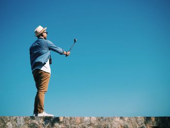 Low angle view of man taking selfie with monopod on stone wall against clear blue sky