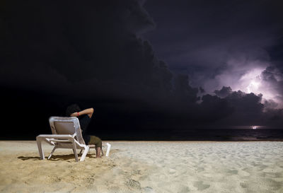 Man sitting on chair at beach against sky at night