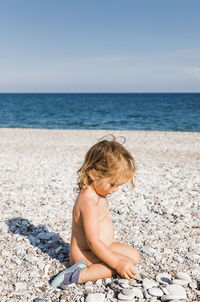 Shirtless girl sitting on pebbles at beach against sky