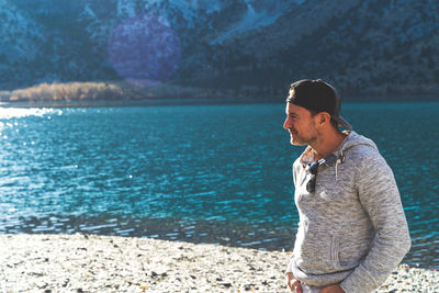 Attractive man standing in front of mountain lake with ball cap on looking away in grey hoodie.
