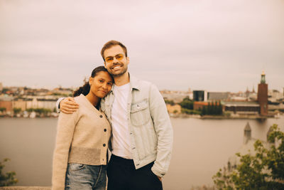 Portrait of male and female friends standing with arm around against lake