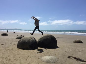 Full length of man leaping on rocks at beach against sky during sunny day