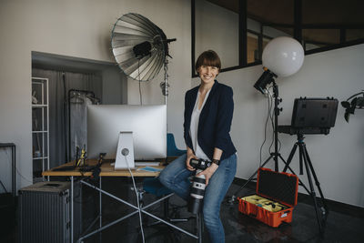 Happy female professional holding camera while sitting on desk at studio