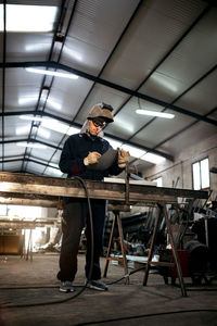 Serious male worker in hardhat and overall taking notes in notebook with pen on table near metal constructions in light garage