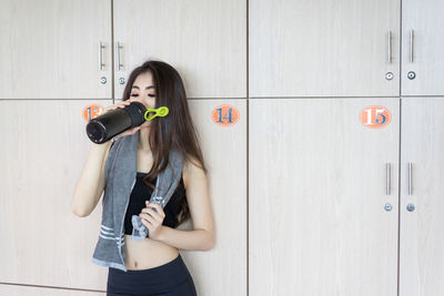 Woman drinking water from bottle while standing against locker