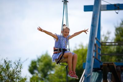 Portrait of happy girl with arms outstretched sitting on swing against sky