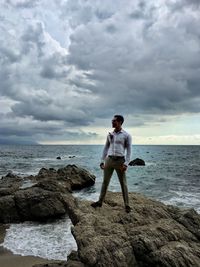 Full length of young man standing on rock at sea shore against sky