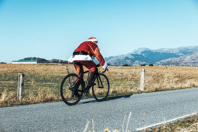 Side view of santa claus in red costume riding modern bike along empty road in highlands on sunny day