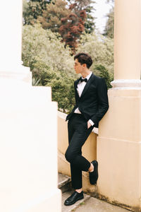 A beautiful young man, the groom in an elegant wedding suit, stands posing in the city's old park