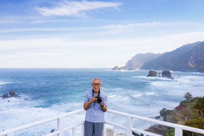 Portrait of man holding camera while standing by railing against sea