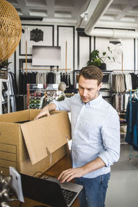 Owner using laptop while unpacking cardboard box in boutique