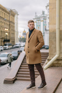 Blond man in beige coat, with dark blue scarf tied around his neck, brown pants and dark shoes