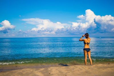 Rear view of woman in bikini standing at seashore against sky