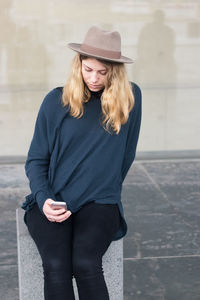 Beautiful young woman using mobile phone while sitting on post against wall