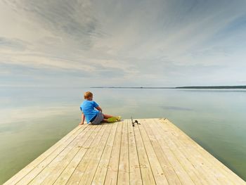 Small fisher on mole. blond boy in blue t-shirt, grey shorts and green flip-flops fishing on pier