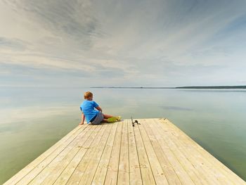 Small fisher on mole. blond boy in blue t-shirt, grey shorts and green flip-flops fishing on pier