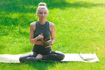 A woman sitting on the green grass in the park in summer, holds a meditation bowl.