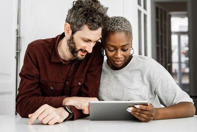 Satisfied african american woman and bearded man smiling while sitting at table and watching digital tablet together at home