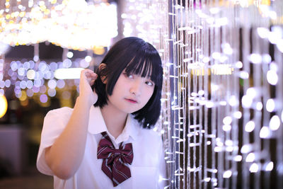 Young woman looking away while standing against illuminated string lights