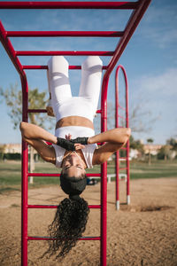 Sportswoman hanging upside down on monkey bars in public park during sunny day