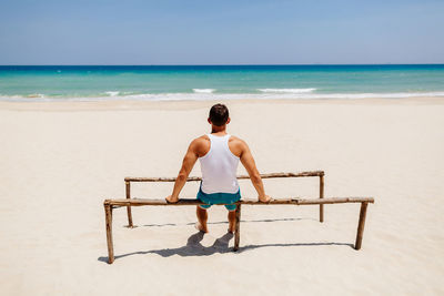 Rear view of man sitting on parallel bars at beach during sunny day