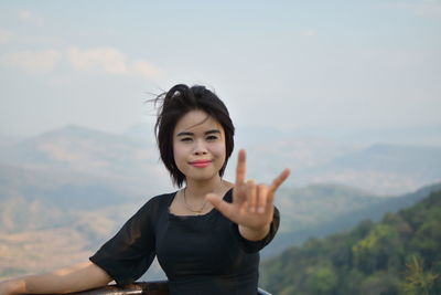 Portrait of smiling young woman gesturing horn sign while standing against mountain and sky
