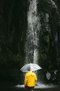 Rear view of man with umbrella standing against waterfall at forest