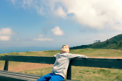 Carefree boy enjoying in sunshine while relaxing with eyes closed on a bench at mountain peak.