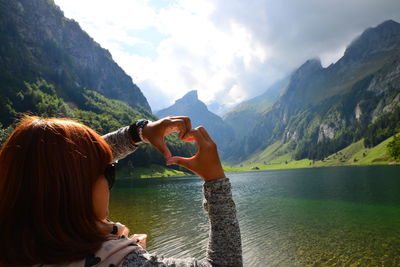 Rear view of woman making heart shape with hands over lake against mountains and sky