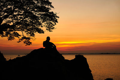 Silhouette man sitting on rock by sea against sky during sunset