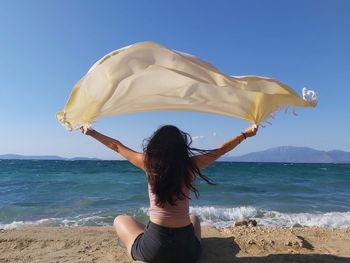 Rear view of woman with arms outstretched holding scarf while sitting at beach against sky