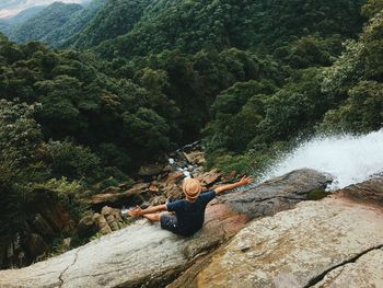 High angle view of man with arms outstretched sitting on rock formation against forest