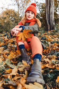 Portrait of cute young woman sitting in autumn leaves wearing knitted warm orange hat and scarf. 