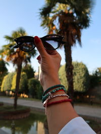 Cropped hand of person holding sunglasses against trees and sky