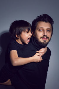 Boy son  sitting on father with a beard and hair back in studio on gray background in black clothes