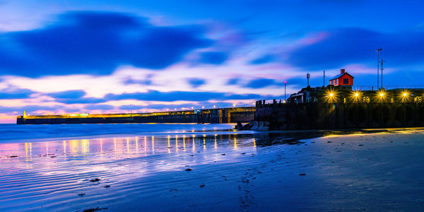 Illuminated building by sea against sky at dusk