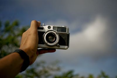 Cropped hand holding camera against sky